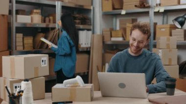 Kızıl saçlı, gözlüklü, dizüstü bilgisayar başında oturan ve Asyalı meslektaşı paketlerle uğraşırken klavyenin detaylarını yazan beyaz bir adam. Küçük işletme kavramı.