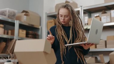 Uzun saçlı, gözlüklü, gözlüklü bir kadın ev ofisinde duruyor ve dizüstü bilgisayarına ciddi bir yüzle paket detaylarını yazıyor. Posta servisi ve küçük işletme kavramı.