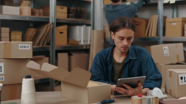 Élégant homme caucasien assis à l'entrepôt et regardant l'ordinateur tablette tout en tapant des détails de colis sur elle. Service postal et concept de petite entreprise. — Video