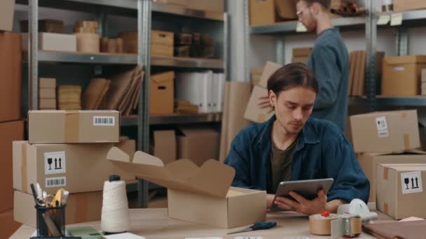 Homme caucasien sérieux assis à la table avec un ordinateur tablette et de travailler attentivement tandis que son collègue masculin boîtes d'emballage à l'arrière-plan. Concept de petite entreprise. — Video