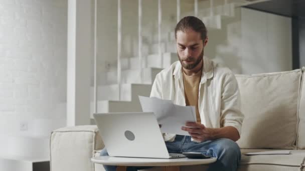 Uomo concentrato seduto sul divano con computer portatile e carte — Video Stock