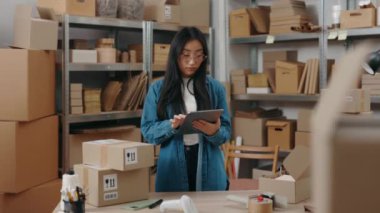 Depoda gözlüklü bir kadın işçi duruyor ve üzerinde paket detaylarını yazarken tablet bilgisayar kullanıyor. Posta servisi ve küçük işletme kavramı.