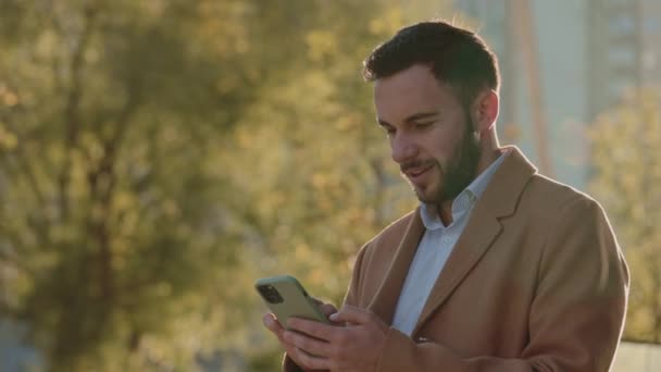 Hombre guapo en abrigo beige leyendo buenas noticias en el móvil — Vídeos de Stock