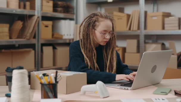 Stylová žena v brýlích sedící ve své kanceláři a s vážnou tváří psala do laptopu podrobnosti o balících. Poštovní služba a koncept malého podnikání. — Stock video