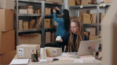 Beyaz tenli, gözlüklü bir kadın dizüstü bilgisayarda oturuyor, paketleri tarıyor ve Asyalı meslektaşı paketleri incelerken klavyede detayları yazıyor. Küçük işletme kavramı.