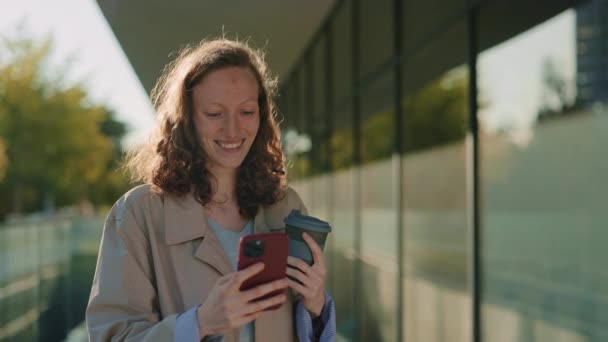 Leende kvinna står utomhus med kaffe och mobil — Stockvideo