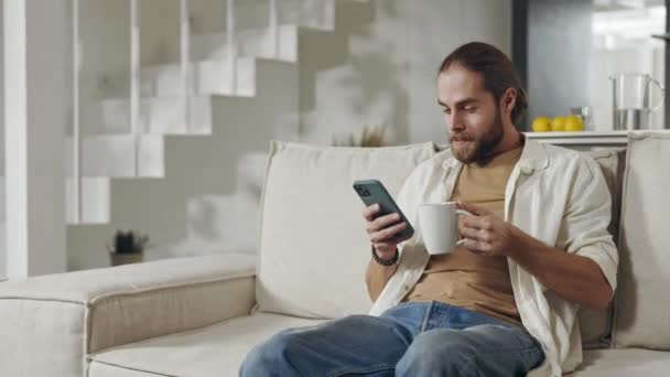 男人喜欢喝热饮和在沙发上使用手机 — 图库视频影像