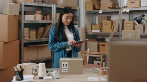 Cintura arriba retrato vista de la tranquila chica asiática morena sosteniendo tableta y paquete de disparos mientras se prepara el envío al cliente. Concepto de pequeña empresa. — Vídeo de stock