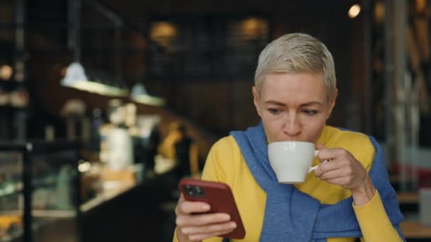 Biała blondynka w średnim wieku z kawą i telefonem w kawiarni — Wideo stockowe
