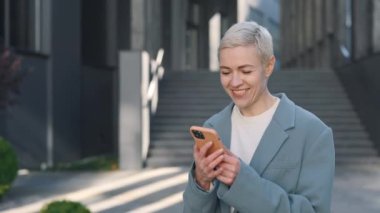 İş merkezinin yakınında yürürken akıllı telefon kullanan mutlu orta yaşlı beyaz kadın. Teknoloji, iletişim, iş konsepti.