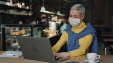 Kafede maskeli bir kadın oturuyor ve dizüstü bilgisayarla yazı yazıyor.