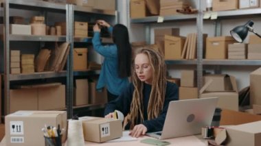 Asyalı esmer meslektaşı paketleri düzenlerken, özenli beyaz kadın dizüstü bilgisayarın başında oturuyor ve paketleri tarıyor. Küçük işletme kavramı,
