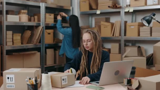 Opmærksom kaukasiske arbejderkvinde sidder på den bærbare computer og scanner pakker, mens hendes asiatiske brunette kollega sorterer gennem pakker. Små og mellemstore virksomheder, – Stock-video