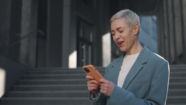 Portrét šťastné obchodní ženy těšit úspěch na smartphone v blízkosti kancelářského centra, Překvapený běloška říká wow, zatímco čtení dobré zprávy na smartphone venku. Úspěch v práci. — Stock video