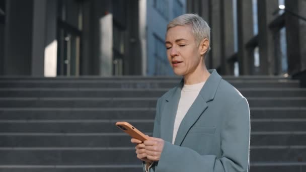 Beautiful middle aged woman in formal wear using modern smartphone looking at camera standing at urban area. Outdoors portrait of smiling lady with short blond hair — Stock Video