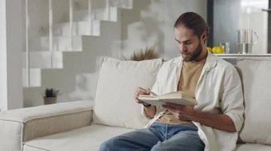 Sakallı adam kanepede oturup kitap okuyor.