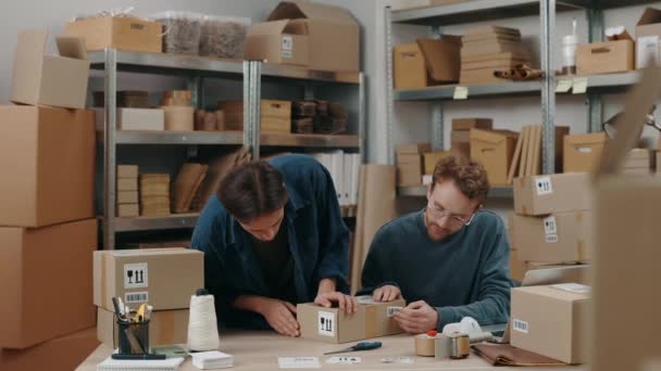 Homme caucasien de gingembre souriant à la caméra avec son collègue masculin tout en emballant des colis à la boîte de placard et la préparation des colis de livraison. Service postal et concept de petite entreprise. Portrait. — Video