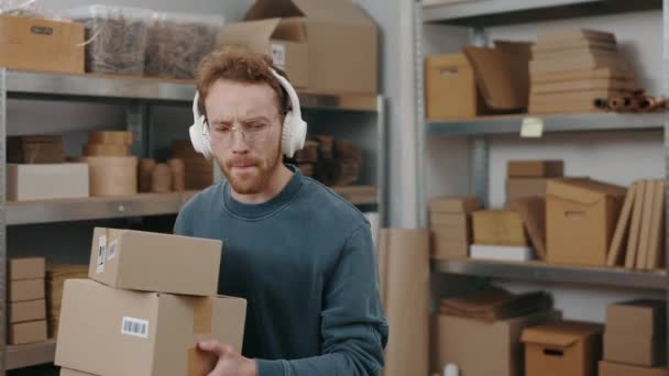 Widok na portret ucieszonego białego mężczyzny noszącego słuchawki słuchającego muzyki podczas noszenia paczek i siedzącego w miejscu pracy z laptopem przy stole. — Wideo stockowe