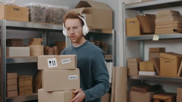 Radosny listonosz noszący słuchawki słuchający muzyki podczas noszenia paczek i siedzący w miejscu pracy z laptopem przy stole. — Wideo stockowe