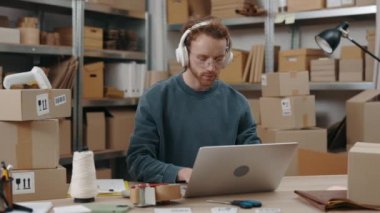 Attentive caucasian man wearing glasses sitting at the laptop computer and typing something at the keyboard while working at the home office. Post service and small business concept.