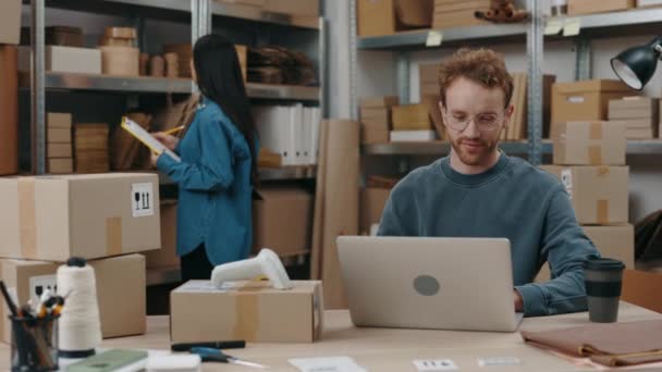 Happy caucasian entrepreneur typing parcels details at the keyboard at the laptop while his female asian colleague sorting through parcels. Small business concept. — Video