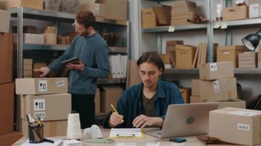 Ginger man standing with tablet at his hands and looking at the shelves while his male colleague working at the laptop at office. Small business concept.