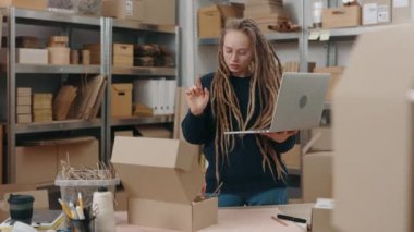 Long haired stylish woman wearing glasses standing at the warehouse and looking at the laptop computer while typing parcel details on it. Post service and small business concept.