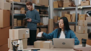 Asyalı şık bir kadın dizüstü bilgisayar başında oturmuş paketleri tarıyor ve beyaz meslektaşı paketleri incelerken ayrıntıları klavyede yazıyor. Küçük işletme kavramı.