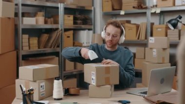 Ginger man wearing headphones sitting at his home office and scanning parcel with serious face while working attentively. Post service and small business concept.