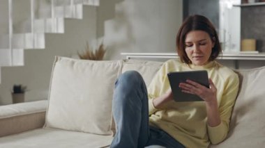 Pleasant woman using digital tablet while resting on couch
