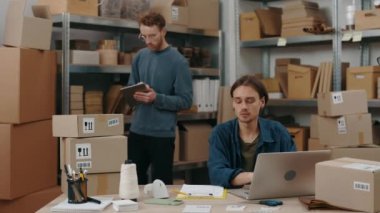 Serious caucasian man typing parcels details at the keyboard at the laptop while his male caucasian colleague sorting through parcels. Small business concept.