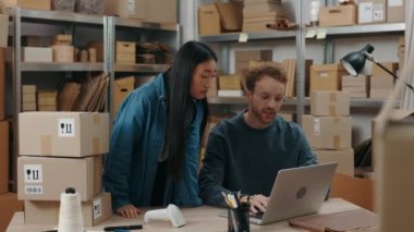 Asian woman putting at the laptop screen with her finger while telling something to her caucasian male colleague while they working at the home office. Small business concept.