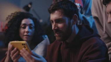 Agitated guy rejoicing while watching over sports betting at the smartphone screen. Focus on the man with device smiling and looking at the friends while sitting on sofa. Sport fans concept.