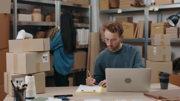 En seriøs, hvit mann som skriver pakker og ser på laptop-skjermen mens hans asiatisk brunette-kollega sorterer gjennom pakker. Småbedriftskonsept. – stockvideo