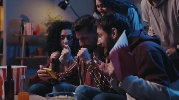 Happy friends rejoicing with real emotions and celebrating winning of favorite team while watching results of the game at the smartphone. Man holding flag and people looking at the screen in awaiting. — Vídeo de Stock