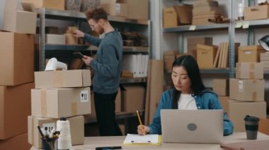 Kafkasyalı erkek arkadaşı paketlerle uğraşırken, Asyalı ciddi bir kadın klavyenin detaylarını diziyor. Küçük işletme kavramı.