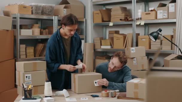 Giving five. Waist up portrait view of the caucasian man and his male colleague putting stickers at the cupboard box while preparing delivery parcel box. Post service and small business concept. — Stok video