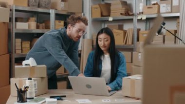 Kızıl Kafkas adam dizüstü bilgisayar ekranına bakıyor ve depoda birlikte çalışırken Asyalı iş arkadaşıyla bazı detayları tartışıyor. Küçük işletme kavramı.