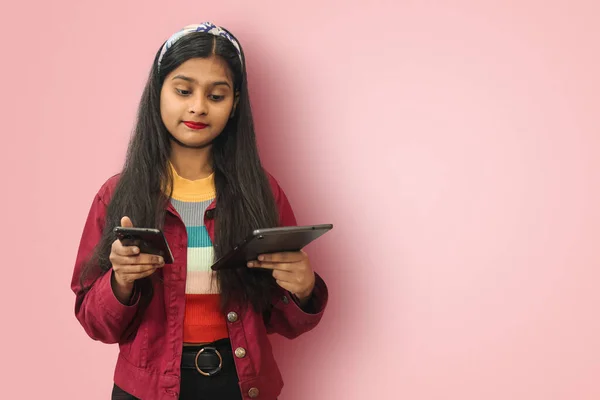Confused Clueless Young Indian Asian teenage girl posing isolated smiling and messaging on mobile phone and tablet using both hands with mockup copy space.
