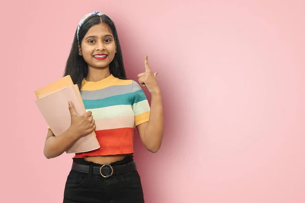 Excited young indian asian student girl posing islolated holding textbooks and pointing up with back right hand, looking directly at camera, dark haired female expressing positive emotions.