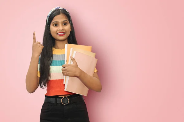 Excited young indian asian student girl posing islolated holding textbooks and pointing up with back left hand, looking directly at camera, dark haired female expressing positive emotions.