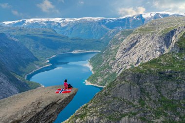 Trolltunga kayası, dağ gölü Ringedalsvatnet manzarası, Norveç. Uçurumda oturan kız Norveç bayrağıyla, 