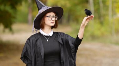 Cadılar Bayramı cadısı. Şapkalı ve cadı kostümlü bir kız portresi ve ormanda siyah bir kuzgun. Tatil cüzdanı ya da hayat için hazırım.