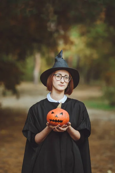 Halloween witch. Portrait of a girl in a hat and a witch costume with a pumpkin in the forest. Ready for the holiday \