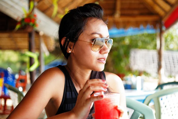 Giovane donna con un top nero e occhiali da sole che si gode un drink in un ristorante sulla spiaggia in Thailandia in una giornata estiva soleggiata — Foto Stock