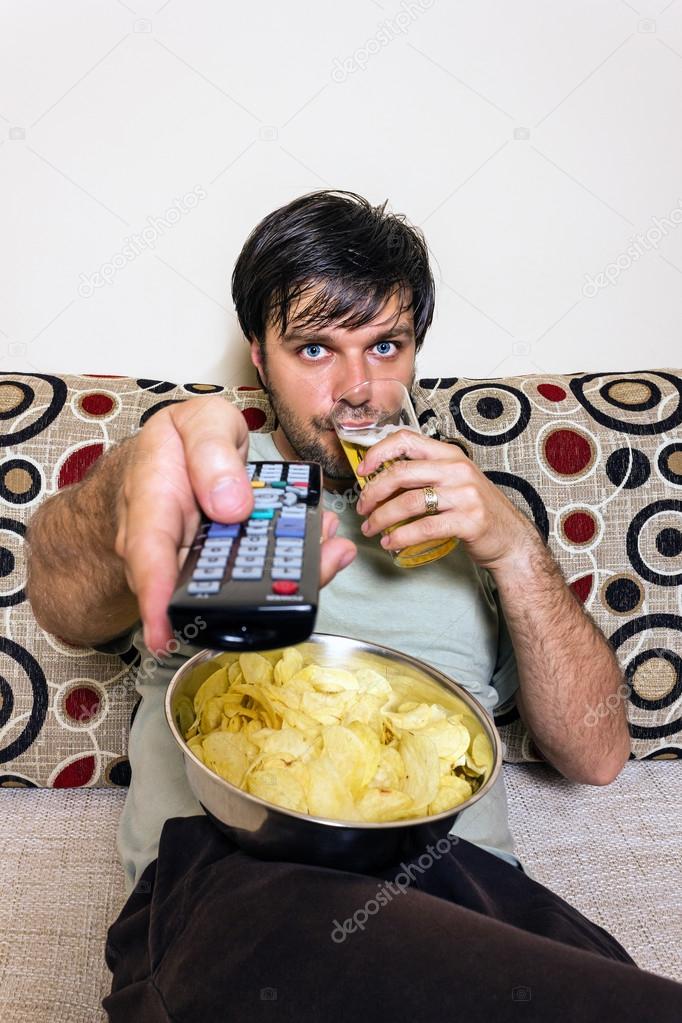 Young man watching television, eating potato chips and drinking