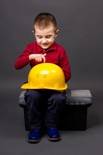 Adorable little boy playing with a helmet while sitting on toolb — Stock Photo, Image