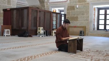 Diz çöküp Kuran okuyan genç adam caminin içindeki ahşap tutacağa farklı açılardan bakıyor camide İslam 'ın kutsal kitabını okuyor.