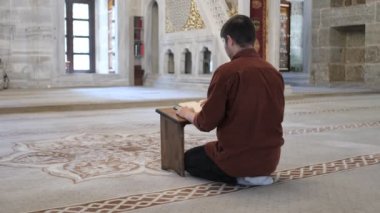 Diz çöküp Kuran okuyan genç adam caminin içindeki ahşap tutacağa farklı açılardan bakıyor camide İslam 'ın kutsal kitabını okuyor.