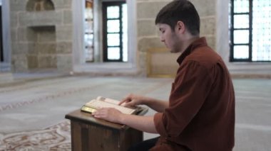 Diz çöküp Kuran okuyan genç adam caminin içindeki ahşap tutacağa farklı açılardan bakıyor camide İslam 'ın kutsal kitabını okuyor.